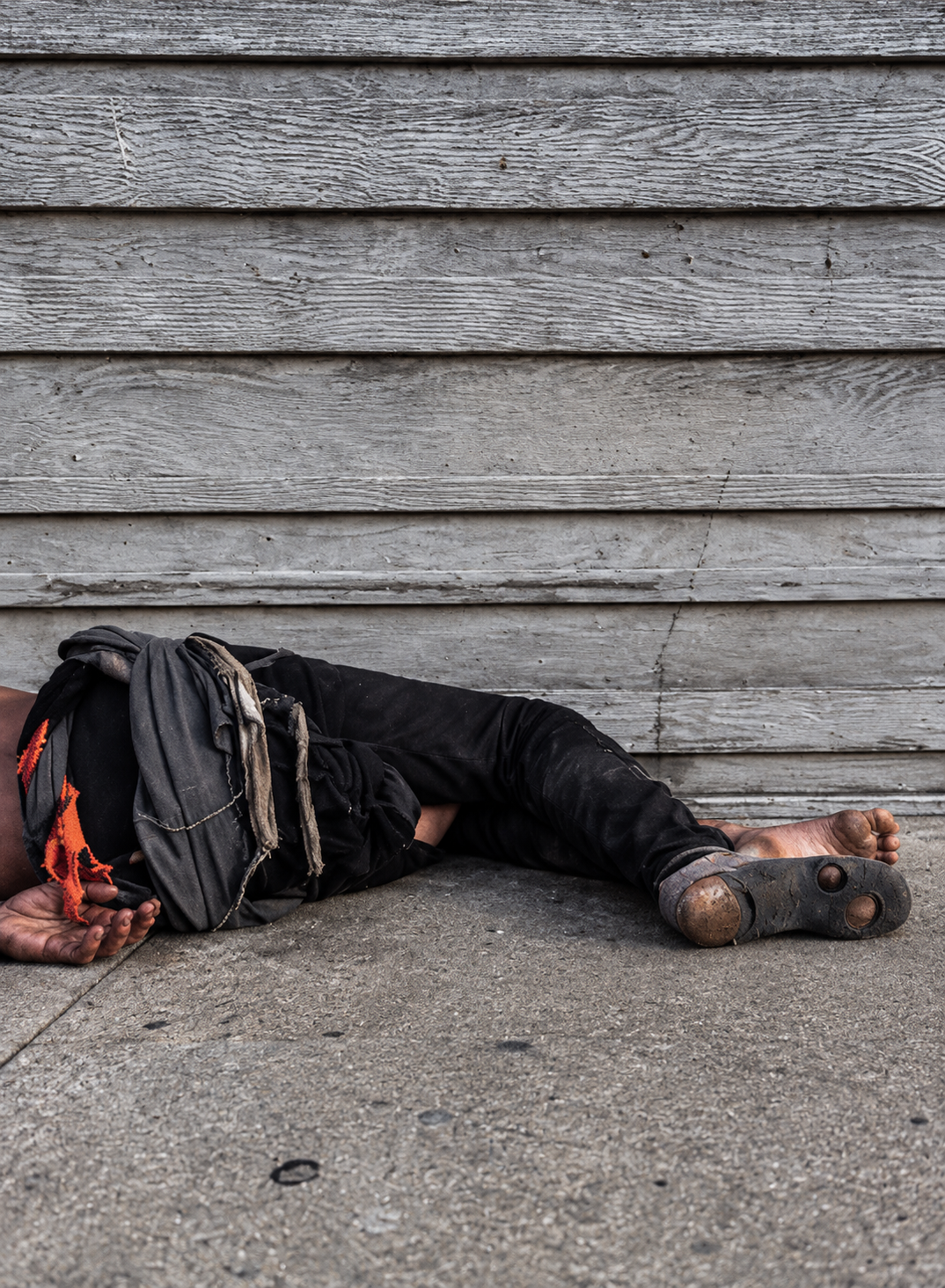 A person lying beside a weathered wooden wall, with bare feet and a worn shoe visible.